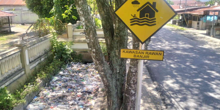 Rambu “Kawasan Rawan Banjir” berdiri tegak di Jalan Cendrawasih, Kota Gorontalo, namun saluran drainase di sisinya justru dipenuhi sampah. Ironi yang mencerminkan masih rendahnya kesadaran lingkungan, sekaligus menjadi ancaman nyata bagi potensi banjir di kawasan tersebut. Foto: Lukman/mimoza.tv