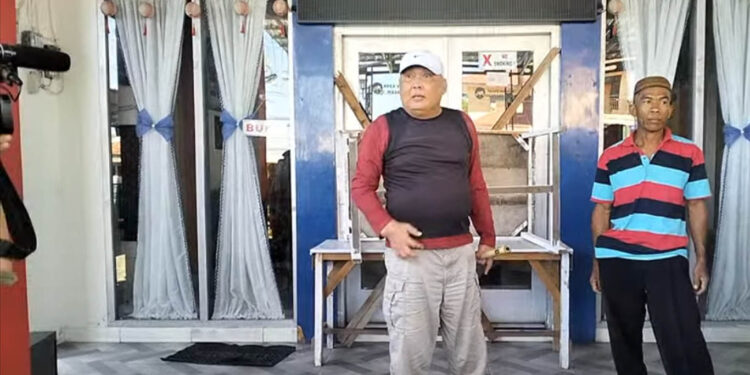 Benny Lengkey (tengah) berdiri di depan pintu Kantor Desa Toto Utara yang telah disegel dalam aksi protes terhadap keputusan pemberhentian sementara Kepala Desa Toto Utara, Ramlah Djafar. Penyegelan mengakibatkan aktivitas di kantor desa terhenti, sementara aparat TNI dan Polri berjaga di lokasi untuk mengamankan situasi.